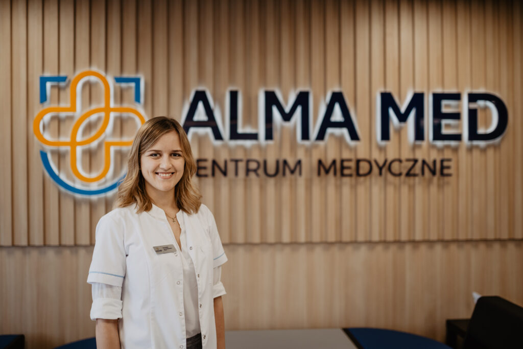 Na zdjęciu lekarz Patrycja Dominika lewandowska, Stoi na pierwszym planie, w tle logo Alma Med. Pomieszczenie to korytarz w przychodni w Boćkach