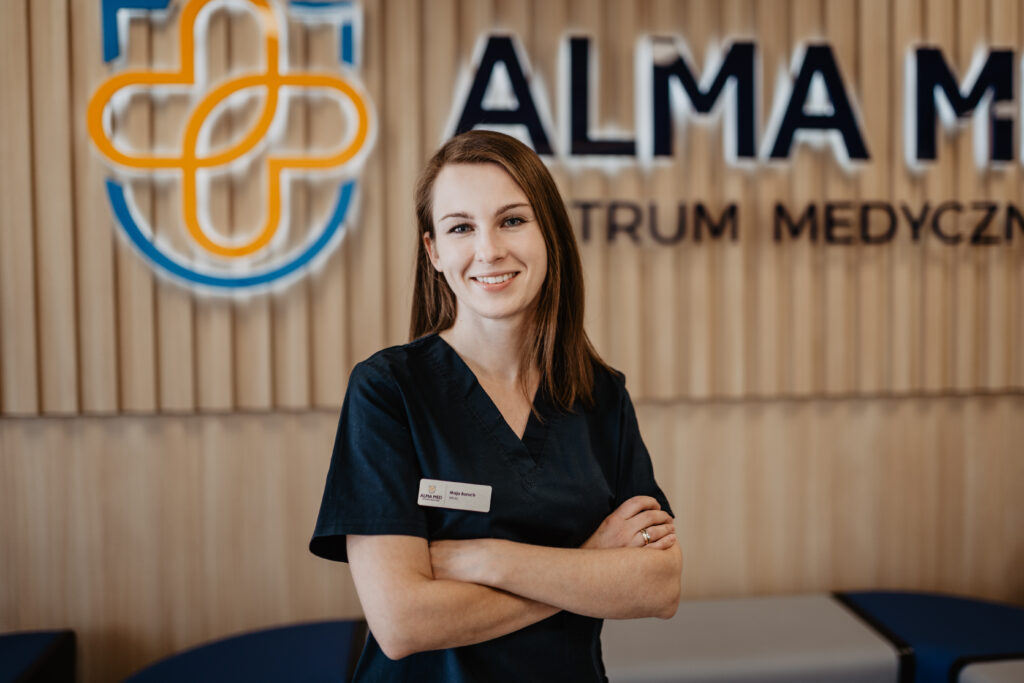Na zdjęciu lekarz Maja Boruch, specjalista medycyny rodzinnej. Stoi na pierwszym planie, w tle logo Alma Med. Pomieszczenie to korytarz w przychodni w Boćkach