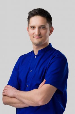Na zdjęciu Michał szamatowicz. Stoi w niebieskiej koszuli na tle białej ściany.
